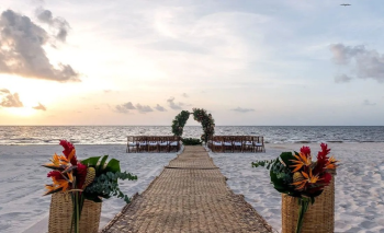 Beach wedding ceremony setup with aisle leading to floral arch overlooking the ocean at sunset.