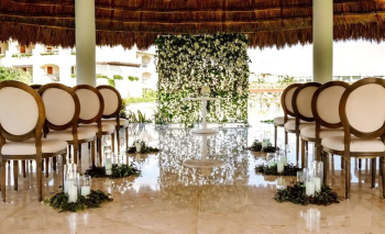 Indoor ceremony setup with aisle, candles, and floral backdrop under a thatched roof gazebo.