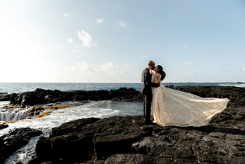 Bride and groom kissing on rocky coastline with ocean waves during destination wedding photos.