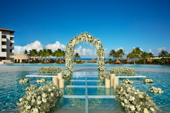 Glass-floor wedding aisle over the pool at the Over The Water venue at Dreams Playa Mujeres, with a white floral arch and ocean views